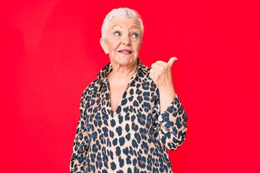 Mavi gözlü, gri saçlı, rahat ve modern leopar desenli bir tişört giyen mutlu yüzüyle gülümseyen ve baş parmağıyla yüzünü gösteren son sınıf güzeli bir kadın.. 