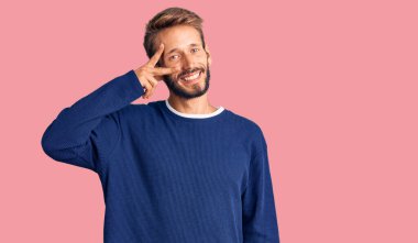 Sakallı yakışıklı sarışın adam, günlük süveter giyiyor, barış sembolü yapıyor, parmakları yüzünde, neşeyle gülümsüyor, zaferini gösteriyor. 