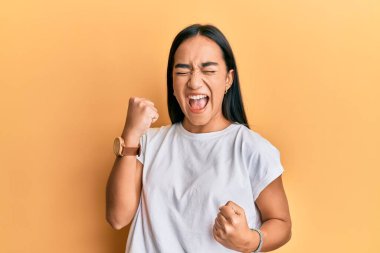 Sıradan beyaz tişört giyen genç Asyalı kadın şaşkınlık ve şaşkınlık içinde kollarını kaldırarak ve gözlerini kapayarak başarıyı kutluyor. 