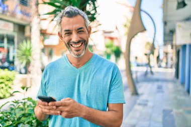 Orta yaşlı, kır saçlı, mutlu gülümseyen bir adam şehrin caddesinde yürürken akıllı telefon kullanıyor..