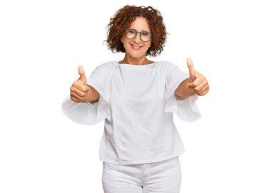 Güzel, orta yaşlı, günlük kıyafetler giyen ve gözlük takan olgun bir kadın. Elleriyle olumlu bir jest yapıyor. Gülümsüyor ve başarı için seviniyor. kazanan jest. 