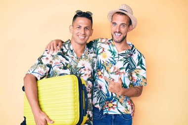 Genç eşcinsel iki adam ellerinde bavullarıyla yaz tatiline gidiyorlar mutlu bir şekilde parmakla gösteriyorlar. 