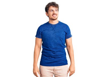 Kıvırcık saçlı, sıradan kıyafetler giyen, yüzünde gülümseme, doğal bir ifade olan genç yakışıklı bir adam. Kendinden emin gülen. 