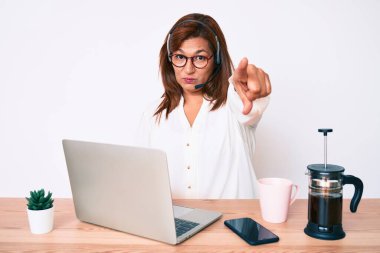 Orta yaşlı esmer, ofiste çalışan, operatör kulaklığı takan, kamerayı işaret eden, kendine güvenen, ciddi bir jest yapan bir kadın. 