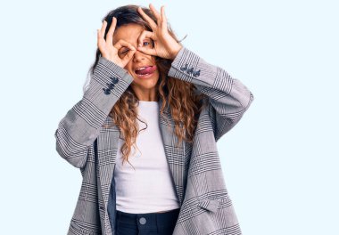 Dövmeli, iş için büyük beden ceket giyen, dilini dışarı çıkaran dürbün gibi jest yapan, parmaklarından bakan Latin kökenli genç bir kadın. çılgın ifade. 