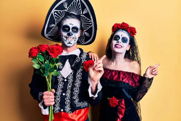 若いですカップル身に着けているメキシコの日の死者の衣装を保持バラ笑顔幸せなポインティングで手と指で側面 — ストック写真