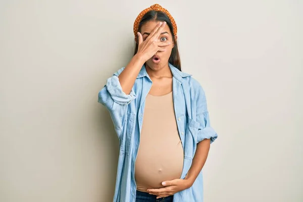 顔と目を手で覆い 恥ずかしい表情で指を通して見て ショックで妊娠中の腹の目に触れて 赤ちゃんを期待美しいヒスパニック系の女性 — ストック写真