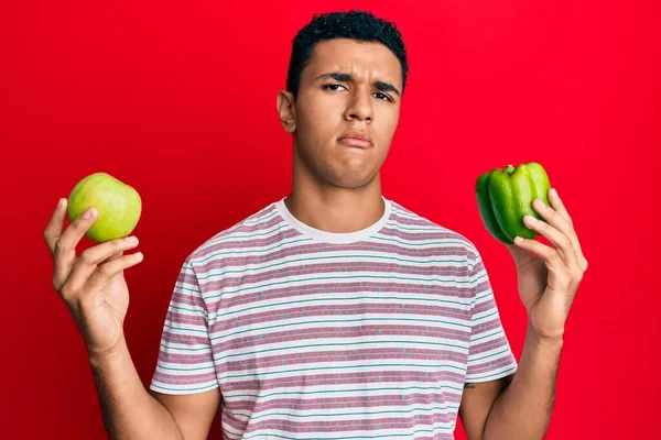 緑のリンゴとコショウを持っている若いアラブ人の男は混乱した表情 疑わしい概念 — ストック写真