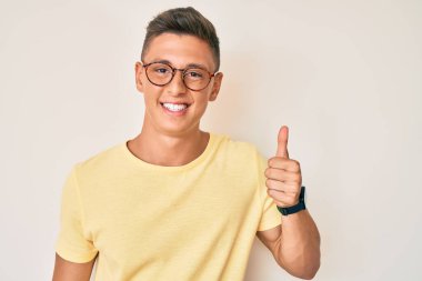 Günlük kıyafetler ve gözlük takan genç İspanyol çocuk mutlu ve pozitif gülümsüyor, başparmağıyla mükemmel ve onay işareti yapıyor. 