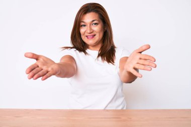 Orta yaşlı esmer, Latin bir kadın. Masada oturmuş kameraya bakıyor. Sarılmak için kollarını açmış gülümsüyor. Mutluluğu kucaklayan neşeli bir ifade.. 