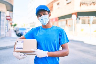 Kurye üniforması giyen Afrikalı kurye dışarıda. Koronavirüs güvenlik maskesi takıyor. Elinde karton bir paket var.