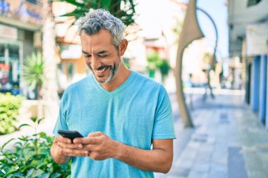 Orta yaşlı, kır saçlı, mutlu gülümseyen bir adam şehrin caddesinde yürürken akıllı telefon kullanıyor..