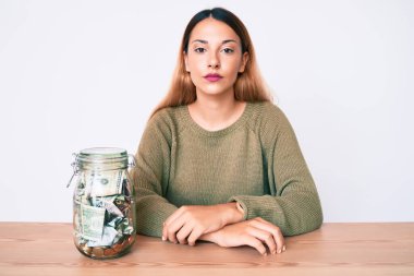 Masada oturan genç esmer kadın. Yüzünde ciddi bir ifade olan bir kavanoz dolusu dolarla. Basit ve doğal bir şekilde kameraya bakıyorsun.. 