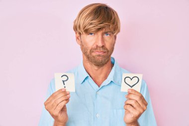 Kalp ve soru işareti taşıyan genç sarışın adam yüzünde ciddi bir ifadeyle rahatladı. Basit ve doğal bir şekilde kameraya bakıyorsun.. 