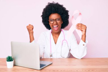 Genç Afrikalı Amerikalı kadın doktor üniforması giyiyor pembe kanser kurdelesi taşıyor gururla bağırıyor, zaferi kutluyor ve başarıyı kollarını kaldırarak kutluyor. 