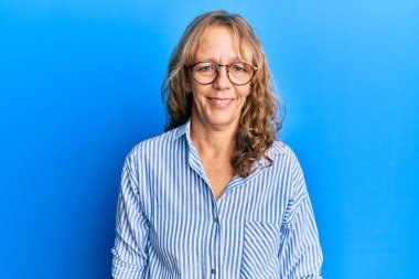 Yüzünde mutlu ve havalı bir gülümsemeyle orta yaşlı, sarışın bir kadın günlük kıyafetler ve gözlük takıyor. şanslı kişi. 