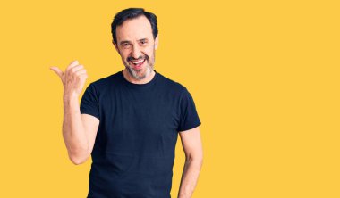 Orta yaşlı yakışıklı, sıradan tişörtler giyen, gülen yüzlü gülümseyen ve baş parmağıyla yan tarafı gösteren bir adam.. 