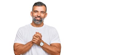 Orta yaşlı yakışıklı bir adam elleri çapraz parmaklı, günlük beyaz tişörtler giyiyor rahat ve neşeli gülümsüyor. Başarı ve iyimserlik 