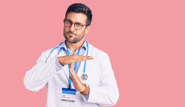 Doktor üniforması ve steteskop giyen genç İspanyol adam. Elleriyle jest yapıyor. Hayal kırıklığına uğramış ve ciddi bir yüz ifadesi var. 