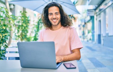 Genç İspanyol gülümseyen adam kahve dükkanının terasında dizüstü bilgisayarla çalışırken mutlu oluyor..