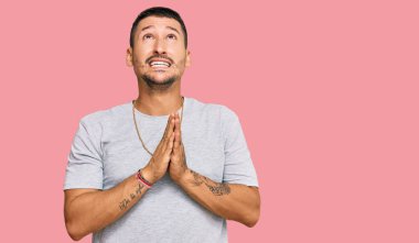 90 'ların tarzında dövmeleri olan yakışıklı bir adam elleri bağlı yalvarıyor ve dua ediyor. Yüzünde umut ifadesi var. Çok duygusal ve endişeli. Yalvarıyor. 