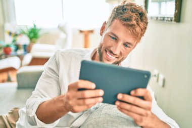 Yakışıklı beyaz adam mutlu bir şekilde evdeki kanepede otururken dokunmatik bir cihaz kullanıyor.