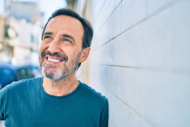 Orta yaşlı, sakallı, mutlu, açık havada duvara yaslanmış bir adam.