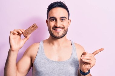 Sakallı, sakallı, enerji proteini yiyen, soyutlanmış pembe arka plan üzerinde gülümseyen, mutlu, eli ve parmağı yanda olan genç bir adam.