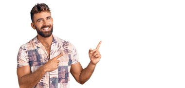 Yaz kıyafetleri giymiş, gülümseyen ve kameraya bakan genç İspanyol adam iki eliyle ve parmaklarıyla kenarı işaret ediyor.. 