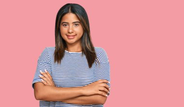 Gündelik kıyafetler giyen, kollarını kavuşturup kameraya gülümseyen genç Latin kız. pozitif kişi. 