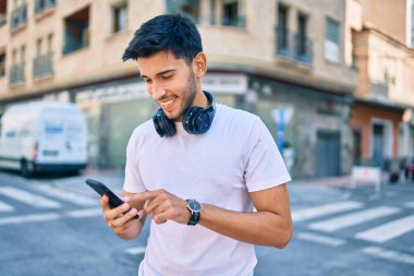 Genç Latin adam şehirde akıllı telefon ve kulaklık kullanarak mutlu bir şekilde gülümsüyor..