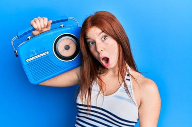 Young red head girl holding vintage radio scared and amazed with open mouth for surprise, disbelief face 