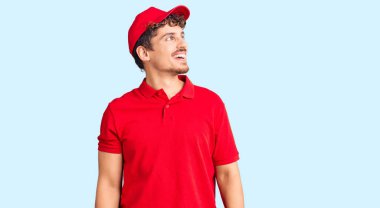 Kıvırcık saçlı, teslimat üniforması giyen, yüzünde gülümseme, doğal bir ifade olan genç yakışıklı bir adam. Kendinden emin gülen. 