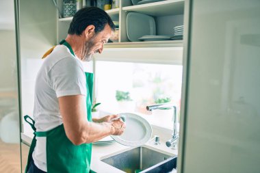 Sakallı, orta yaşlı bir adam mutlu bir şekilde evde bulaşık yıkıyor.
