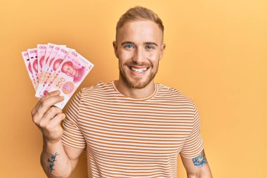 Young caucasian man holding 100 yuan chinese banknotes looking positive and happy standing and smiling with a confident smile showing teeth 