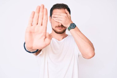 Sıradan beyaz tişört giyen genç yakışıklı adam elleriyle gözlerini kapatıyor ve üzgün ve korku ifadeleriyle jest yapıyor. utangaç ve olumsuz kavram. 