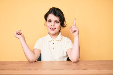 Gündelik giysiler giyen tatlı İspanyol çocuk masaya oturmuş başarılı bir fikirle işaret ediyor. Heyecanlı ve mutlu. Bir numara.. 