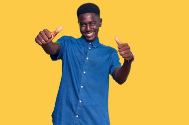 Genç Afro-Amerikan adam günlük giysiler giyip olumlu jestler yapıyor. Elleriyle gülümsüyor ve başarı için seviniyor. kazanan jest. 