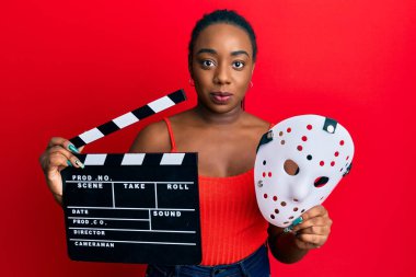 Genç Afro-Amerikalı kadın elinde video film klasörü ve korku maskesi tutarken yüzünde ciddi bir ifadeyle rahatladı. Basit ve doğal bir şekilde kameraya bakıyorsun.. 
