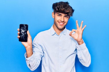 Kırık akıllı telefon tutan genç İspanyol adam kırık ekranı gösteriyor tamam işareti yapıyor parmaklarıyla, gülümseyerek mükemmel bir sembol oluşturuyor. 
