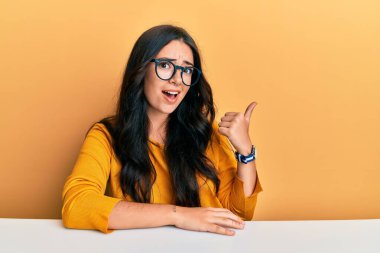 Güzel, esmer, gözlüklü, sıradan elbiseli bir kadın masaya oturmuş gülümsüyor ve başparmağıyla yan tarafa bakıyor.. 