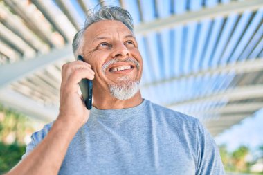 Orta yaşlı, ispanyol, kır saçlı, gülümseyen, mutlu, parkta akıllı telefondan konuşan bir adam..