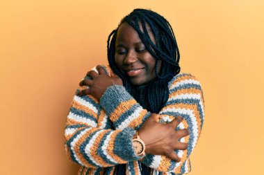 Saçları örgülü genç siyahi bir kadın sıradan bir kazak giyiyor mutlu ve pozitif, kendinden emin bir şekilde kendine sarılıyor. Sevgi ve özgüven. 