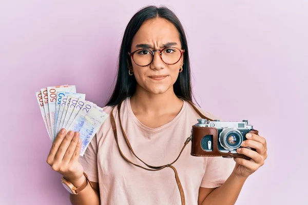 Young asian woman holding vintage camera and swedish krona skeptic and nervous, frowning upset because of problem. negative person.