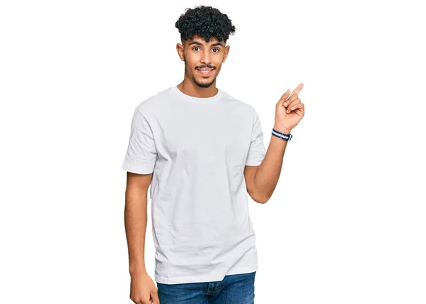 年轻的阿拉伯男子 身穿宽松的白色T恤 脸上挂着大大的笑容 手指头指向侧面看相机 — 图库照片