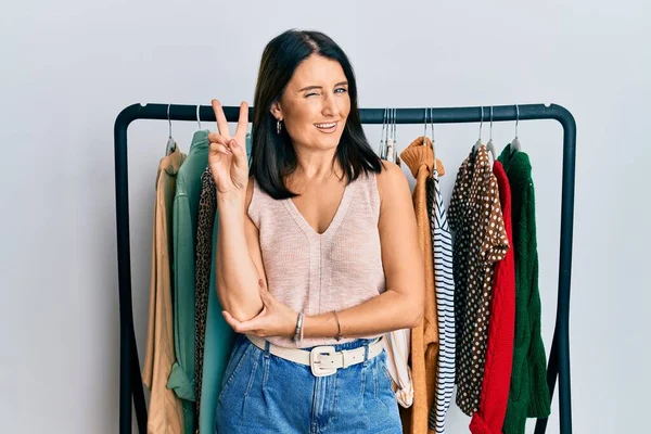 Middle age brunette woman working as professional personal shopper smiling with happy face winking at the camera doing victory sign. number two.