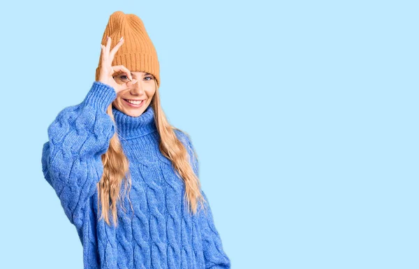 美しいですCaucasian女性とともにブロンド髪身に着けているウールセーターと冬の帽子笑顔幸せな行いますOk Signとともに手で目を通して指を見て — ストック写真