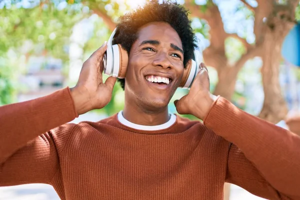 ヘッドフォンを使用して音楽を聴く若いハンサムなアフリカ系アメリカ人男性 町の通りを歩いていると笑顔で立って — ストック写真