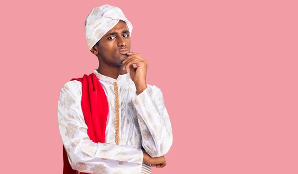 伝統的なシャーワニを身に着けているアフリカのハンサムな男は 集中的な表現について考える顎に手で服を見た 思慮深い顔で笑顔 疑わしい概念 — ストック写真