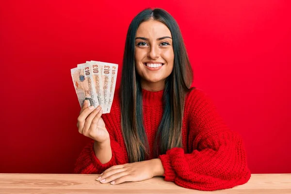 Beautiful hispanic woman holding united kingdom pounds looking positive and happy standing and smiling with a confident smile showing teeth